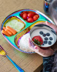 Child enjoying healthy meal on stainless steel rainbow plate with fruits, vegetables, rice, and colorful fork.
