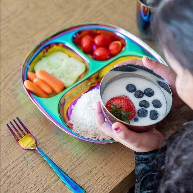 Child enjoying healthy meal on stainless steel rainbow plate with fruits, vegetables, rice, and colorful fork.